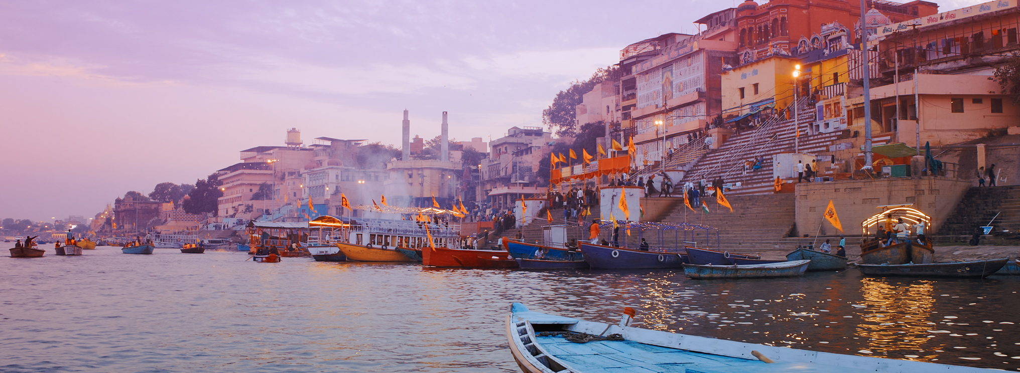 Ein Fluss bei Sonnenaufgang, mit Booten und Stadt am Ufer – die Szene spiegelt die spirituelle Bedeutung des Wassers und den ewigen Kreislauf von Leben und Tod im Hinduismus wider.