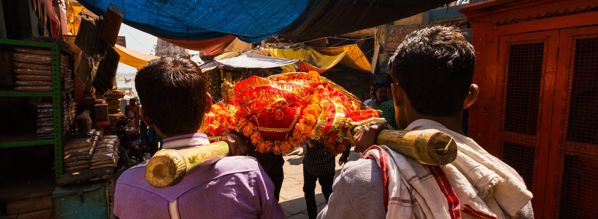 Zwei Männer tragen eine Bahre durch eine enge Gasse. Die Szene verweist auf hinduistische Bestattungsrituale und den Weg vom Leben in den Tod als Teil eines spirituellen Kreislaufs.