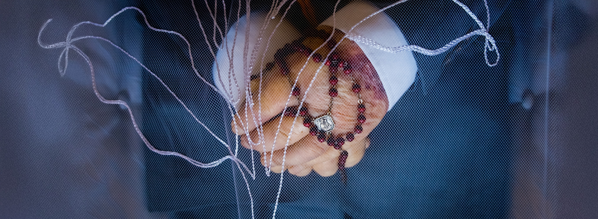 Hinter einem Schleier ruhen zwei gefaltete Hände, auf denen ein Rosenkranz liegt. Die Szene verweist auf stille Trauer, Glaube und Gebet.