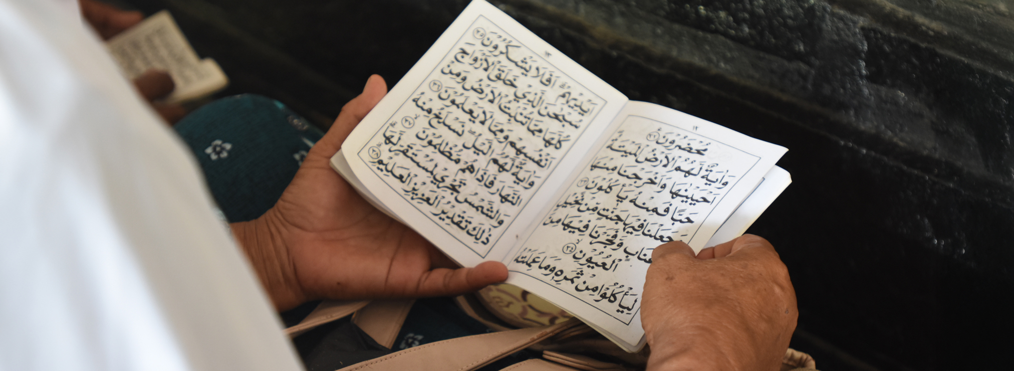 Hände halten ein geöffnetes Buch mit arabischer Schrift. Die Szene zeigt persönliche Koranrezitation und spirituelle Nähe im Angesicht des Todes.